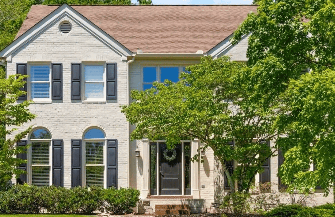 Two-story light brick house with dark shutters and arched windows, partly obscured by a leafy tree.