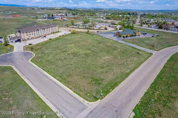 Aerial view of a vacant grassy lot at a road intersection, with a multi-unit building nearby.