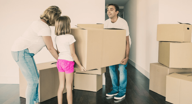 A family carries a large cardboard box through a home filled with moving boxes.