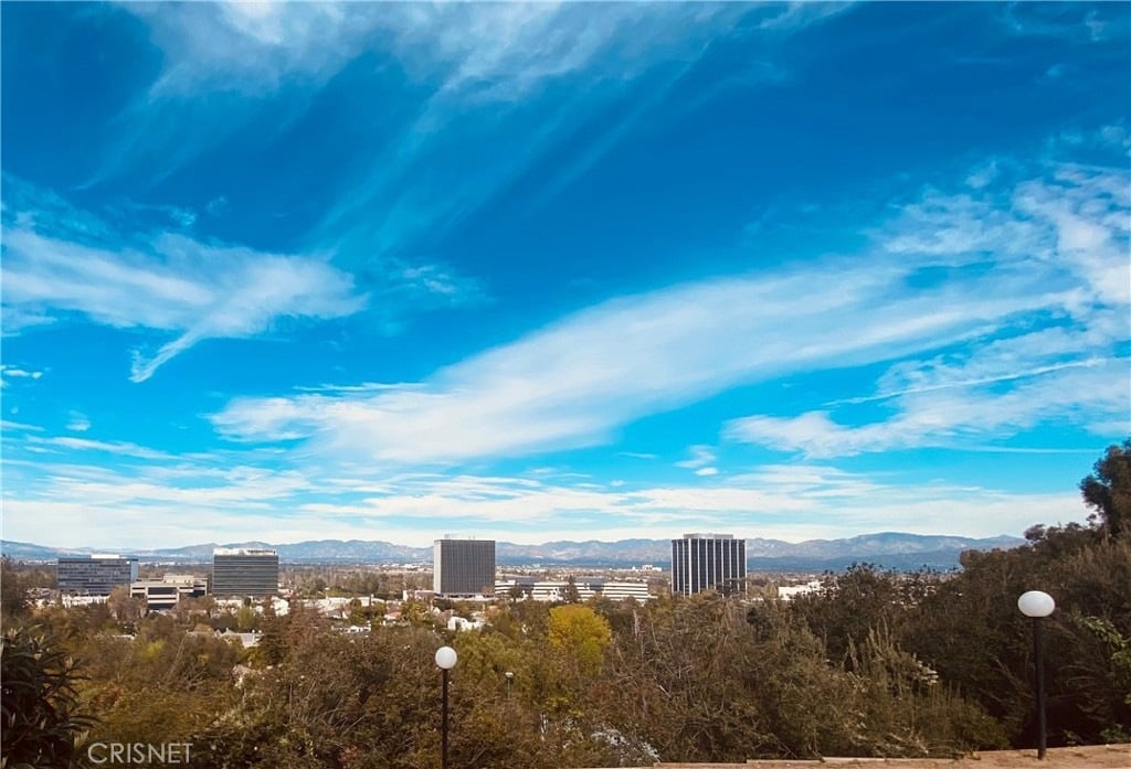 Mountain range and office buildings seen beyond trees beneath a wide, cloud-streaked blue sky.