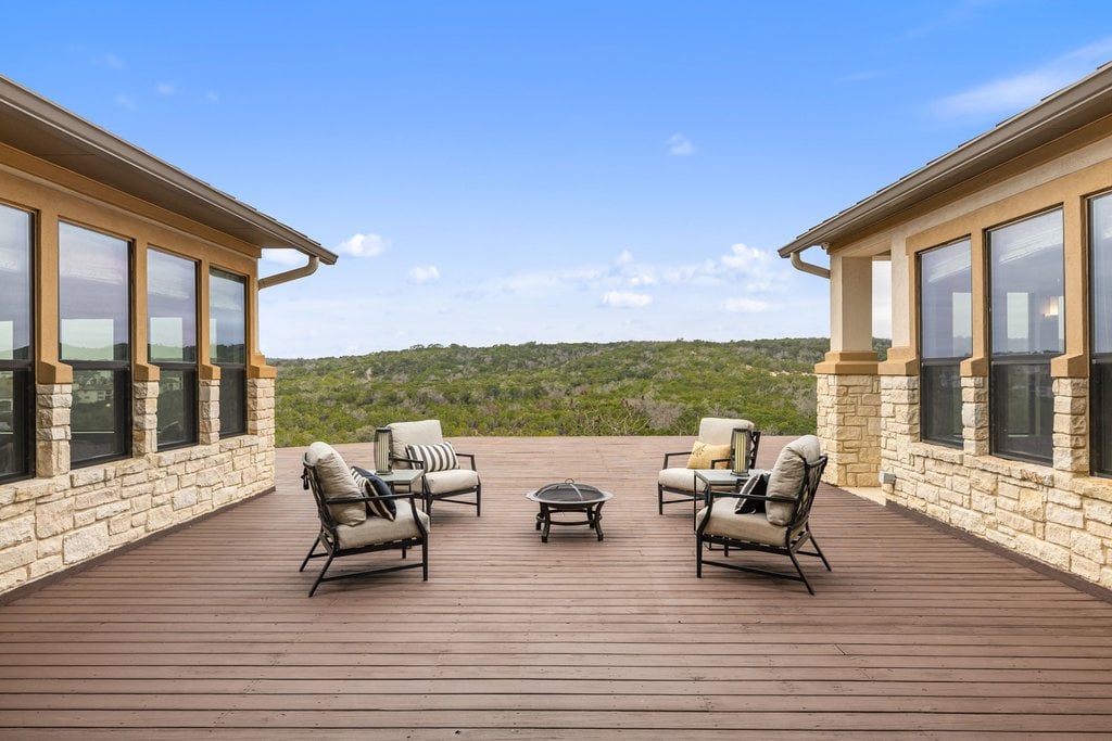 Furnished wood deck between stone-sided home wings overlooking a wooded hillside.
