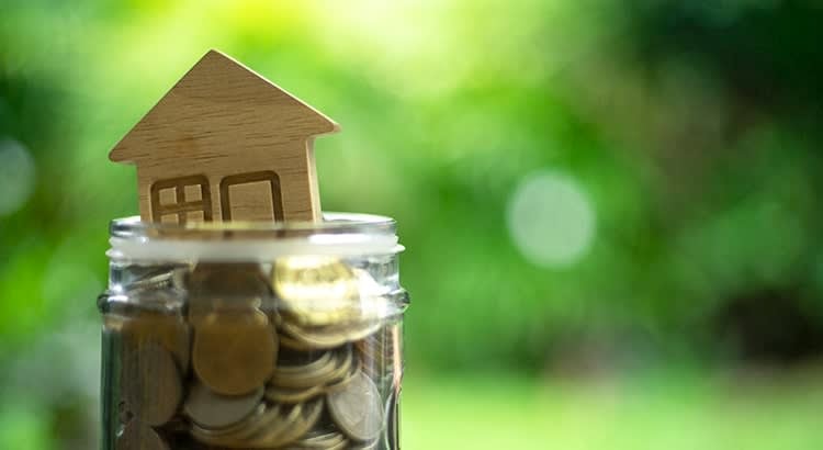 A miniature wooden house sits on top of a jar filled with coins, representing savings or investment in real estate, possibly linked to concepts of home ownership or financial planning.