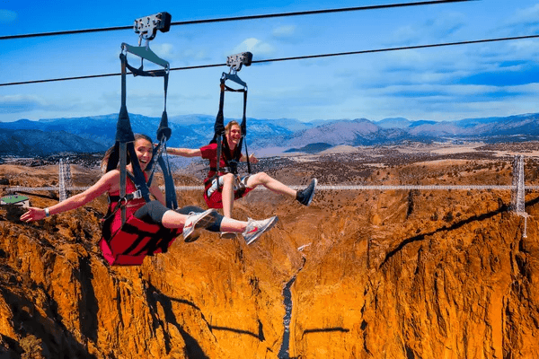 Two people zipline above an orange-rock canyon with distant mountains.