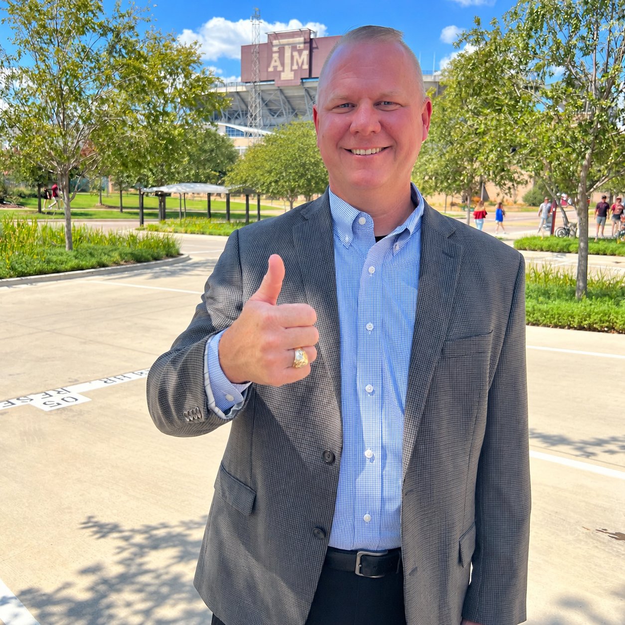 Man in a gray blazer gives a thumbs-up in front of an A&M stadium.