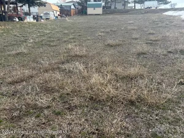 Grassy lot with sparse snow and buildings along its far edge.