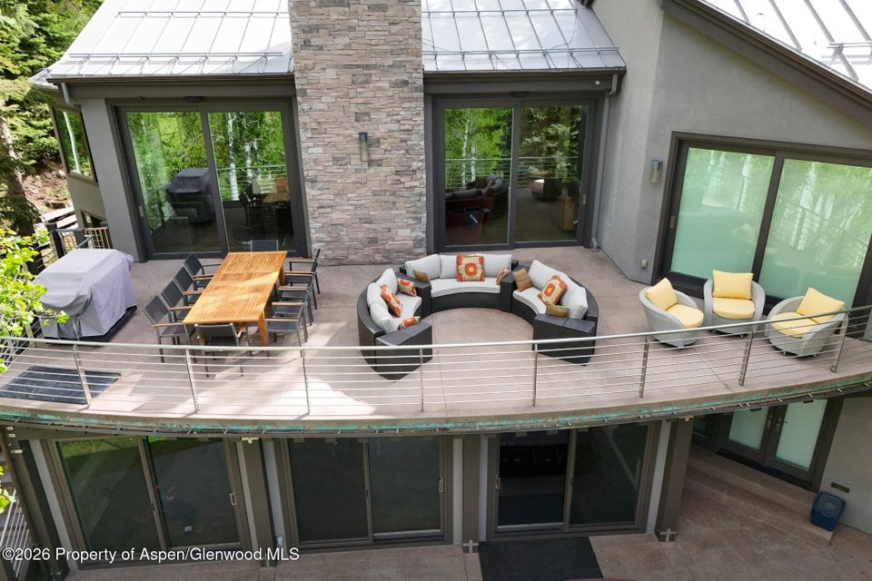 Furnished patio with curved sofa, yellow lounge chairs, and wooden dining table beside a stone-clad home.