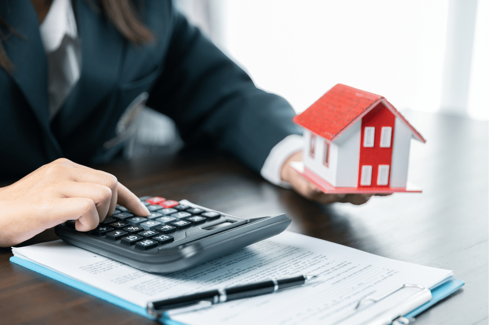 Person calculates real estate paperwork while holding a red-and-white model house.