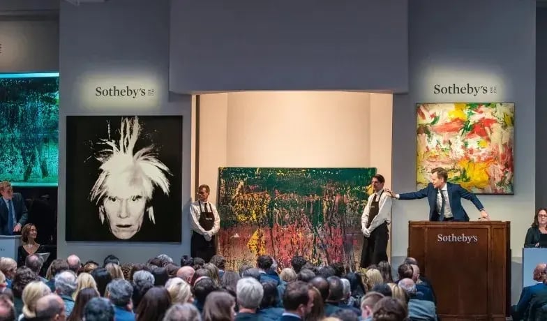 An auction room filled with a large audience seated, facing a stage where an auctioneer stands behind a wooden podium with "Sotheby's" inscribed on it.
