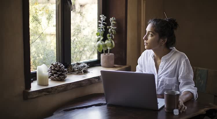 A woman sits at a table with a laptop, gazing out the window. The setting suggests a quiet moment of reflection, possibly while working from home.