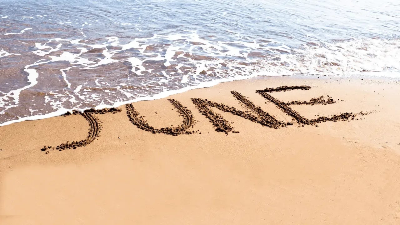 The word "June" written in the sand on a beach with waves approaching, symbolizing the summer month and a sense of relaxation or vacation.