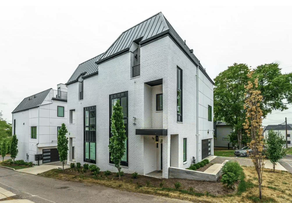Modern white-brick home with a steep dark metal roof, tall windows, and a landscaped front yard.