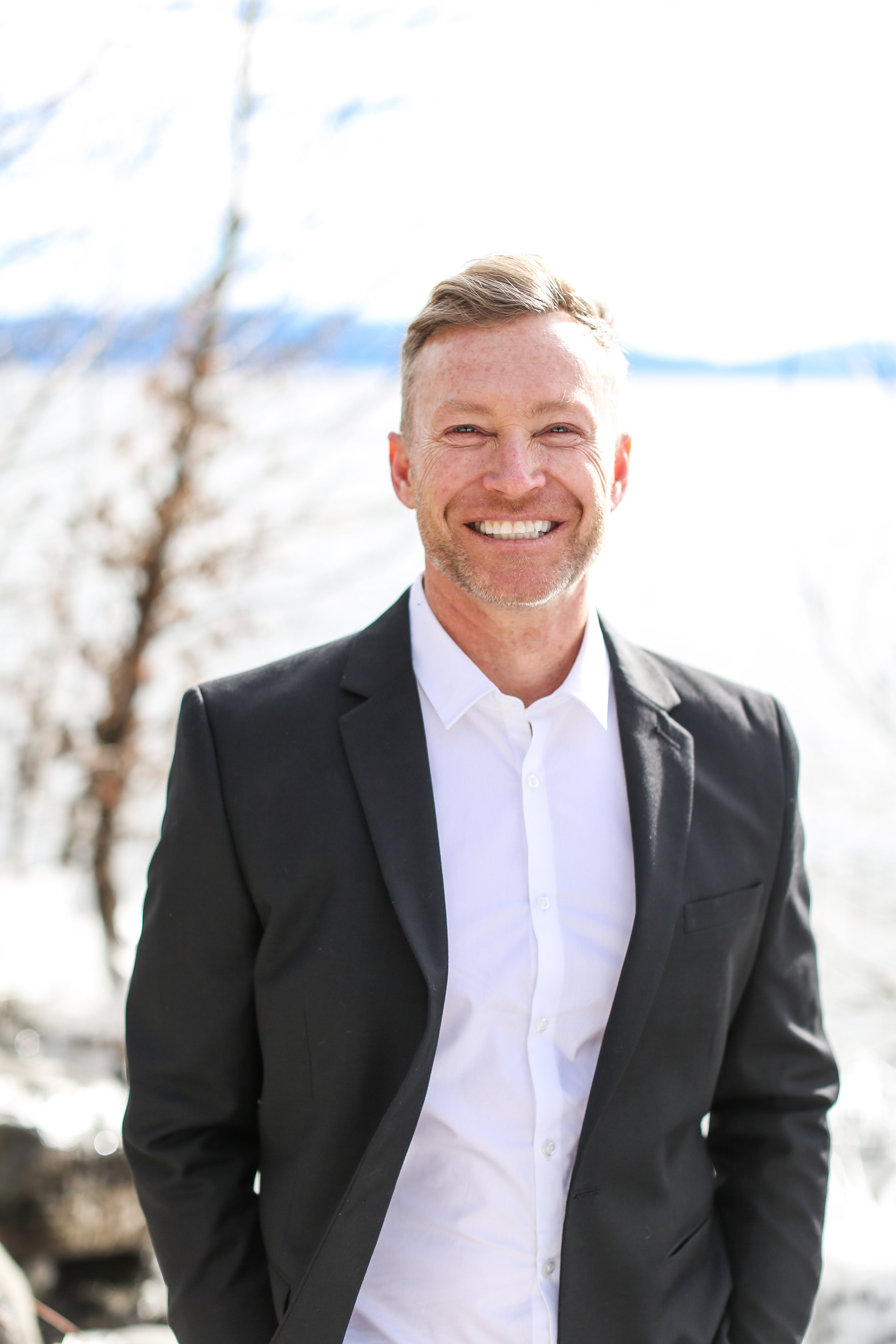 Smiling man in a black suit and white shirt outdoors against a snowy landscape.