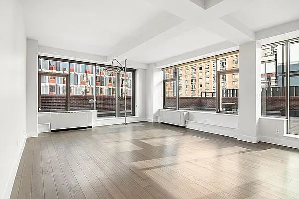 Vacant apartment living room with wide windows, light wood floors, and exposed ceiling beams.