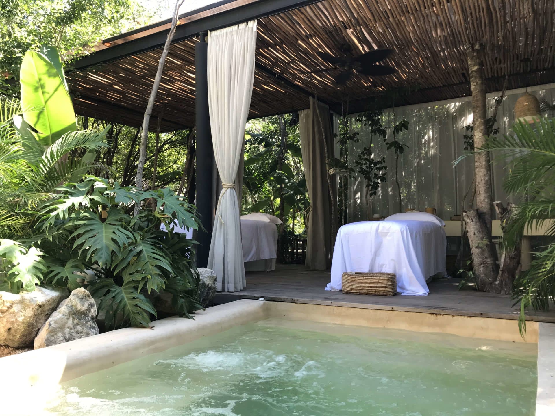 Outdoor pool beside a thatched, curtained cabana surrounded by tropical plants.