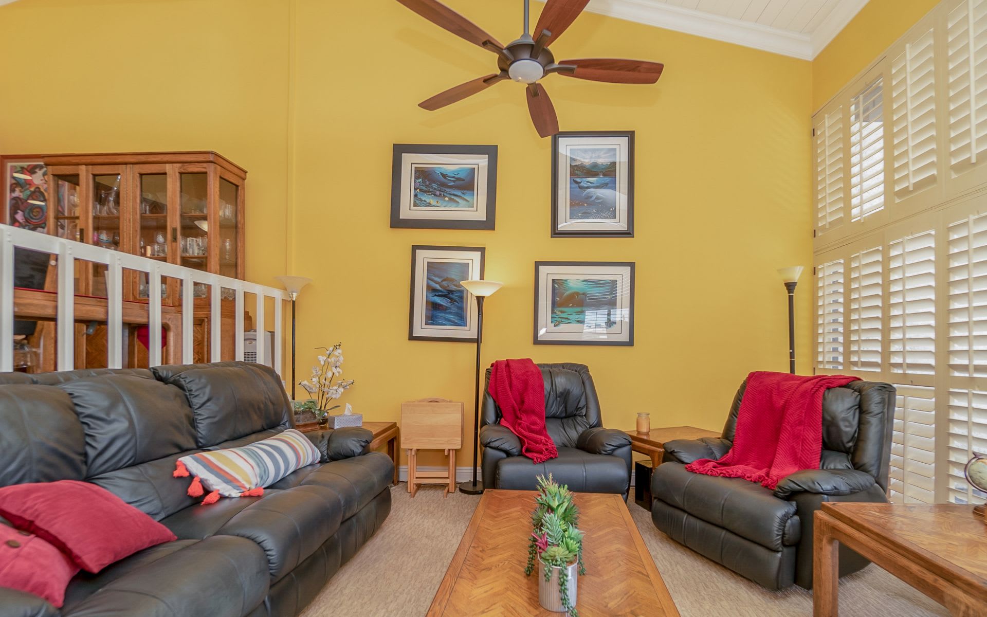 Living room with yellow walls, black leather seating, red throws, and white plantation shutters.