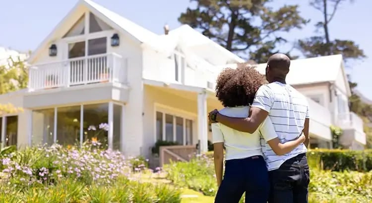  A couple standing in front of a large, modern house, with their arms around each other. They appear to be admiring their home or discussing their future.