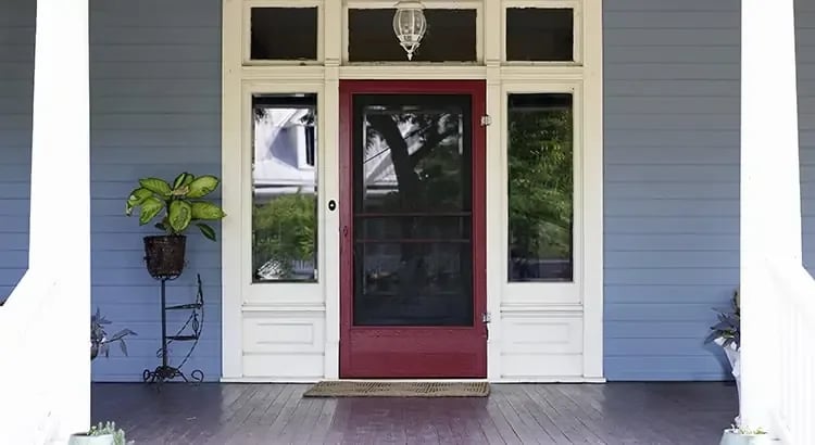 A front porch with a red door, flanked by potted plants. The exterior of the house is painted in a light blue shade, giving it a welcoming appearance.