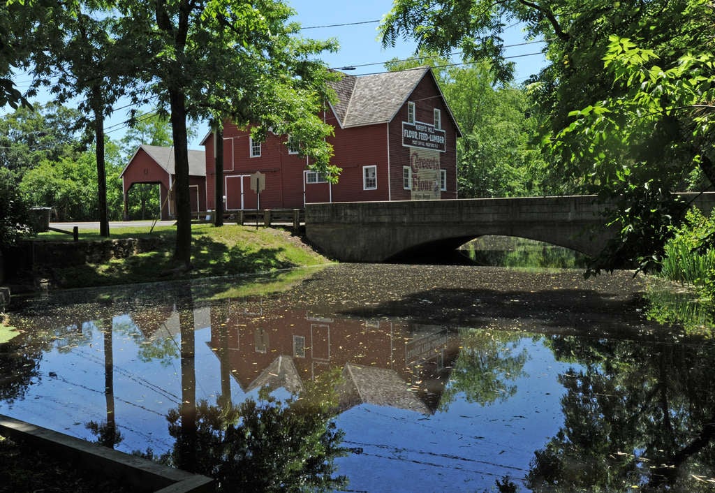 Red wooden mill and stone bridge reflected in a tree-lined pond.