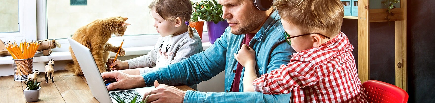 A father works on a laptop as two children draw beside him and a ginger cat sits on the table.