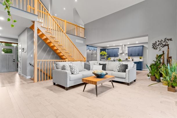 Open-concept living room with a gray sectional, blue kitchen cabinets, and a wooden staircase.