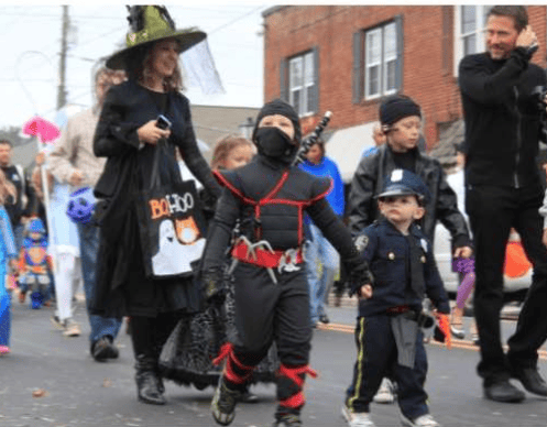 Children in superhero and police costumes walk in a Halloween parade beside an adult dressed as a witch.