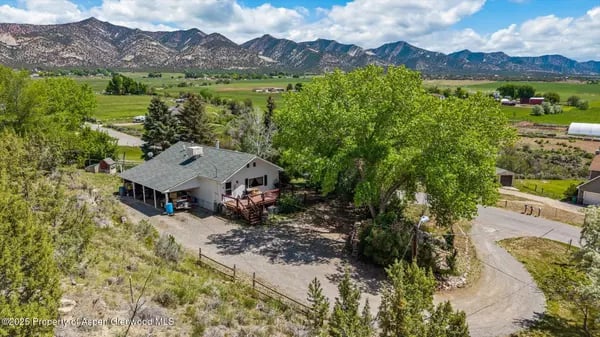 Rural home with mature trees, a broad driveway, open fields, and mountains beyond.