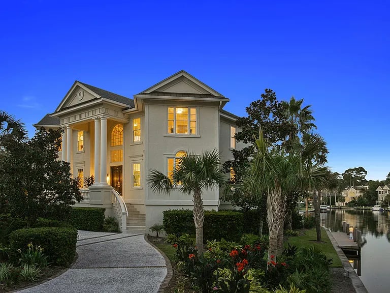Two-story home with a columned entrance and lit windows beside a canal at dusk.