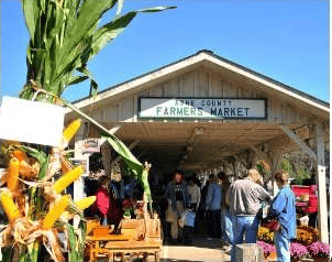 Farmers market beneath a wooden pavilion, with shoppers and produce at the entrance.