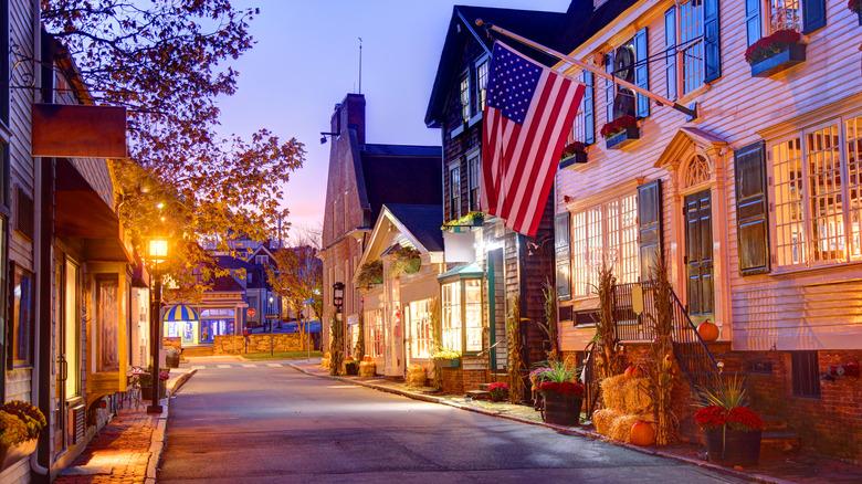 Historic storefronts line a warmly lit street at dusk, with an American flag and autumn decorations.