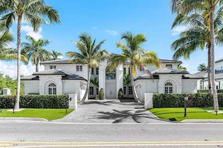 White two-story residence with arched windows, a broad driveway, clipped hedges, and tall palms.