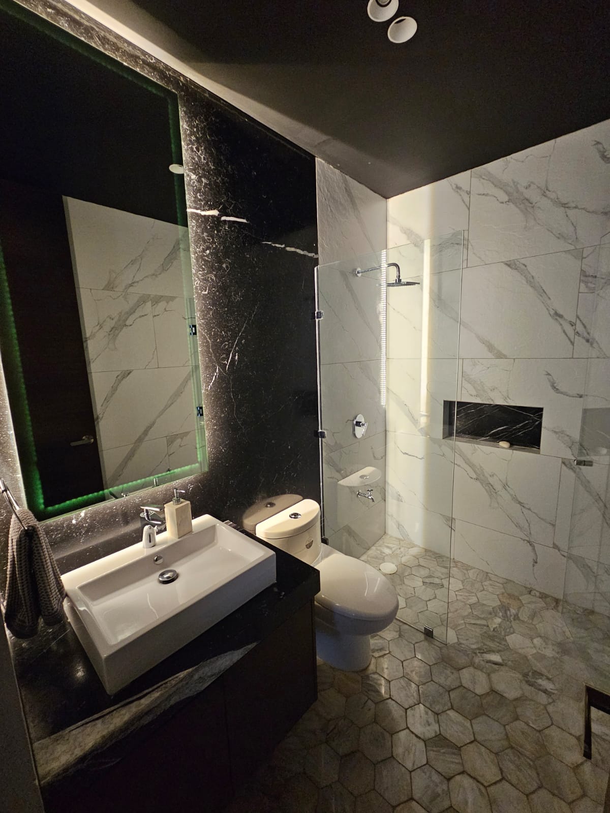 Modern bathroom with a glass shower, marble-look walls, dark stone vanity, and hexagonal floor tiles.