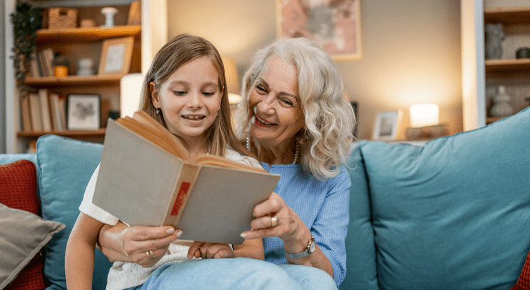 Grandmother and granddaughter reading together on couch – multigenerational family bonding in cozy home setting.