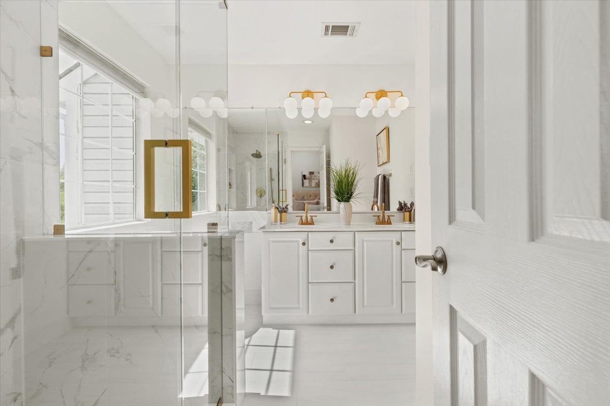 Bright white bathroom with a glass shower, double vanity, marble-look tile, and gold fixtures.