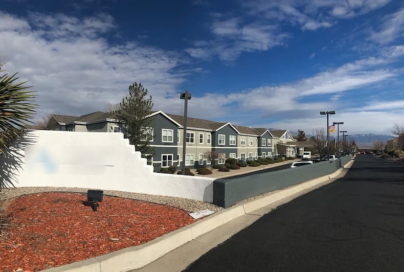 Two-story senior living community behind a stucco entrance wall, with distant mountains.