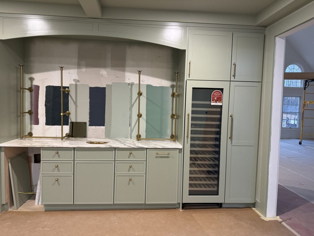 Renovation kitchen with pale green cabinets, brass hardware, and paint samples on the unfinished backsplash.