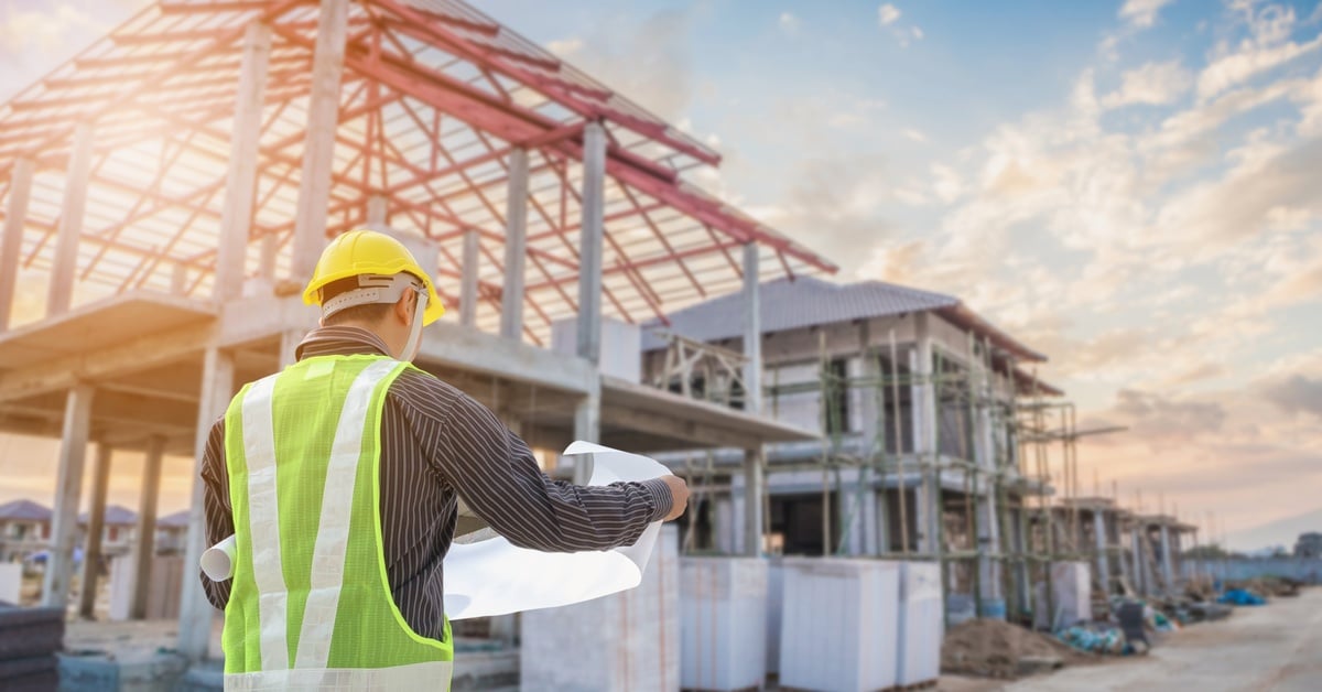 A rear view of a contractor in a high-visibility vest and hard hat examining blueprints in front of an unfinished house.