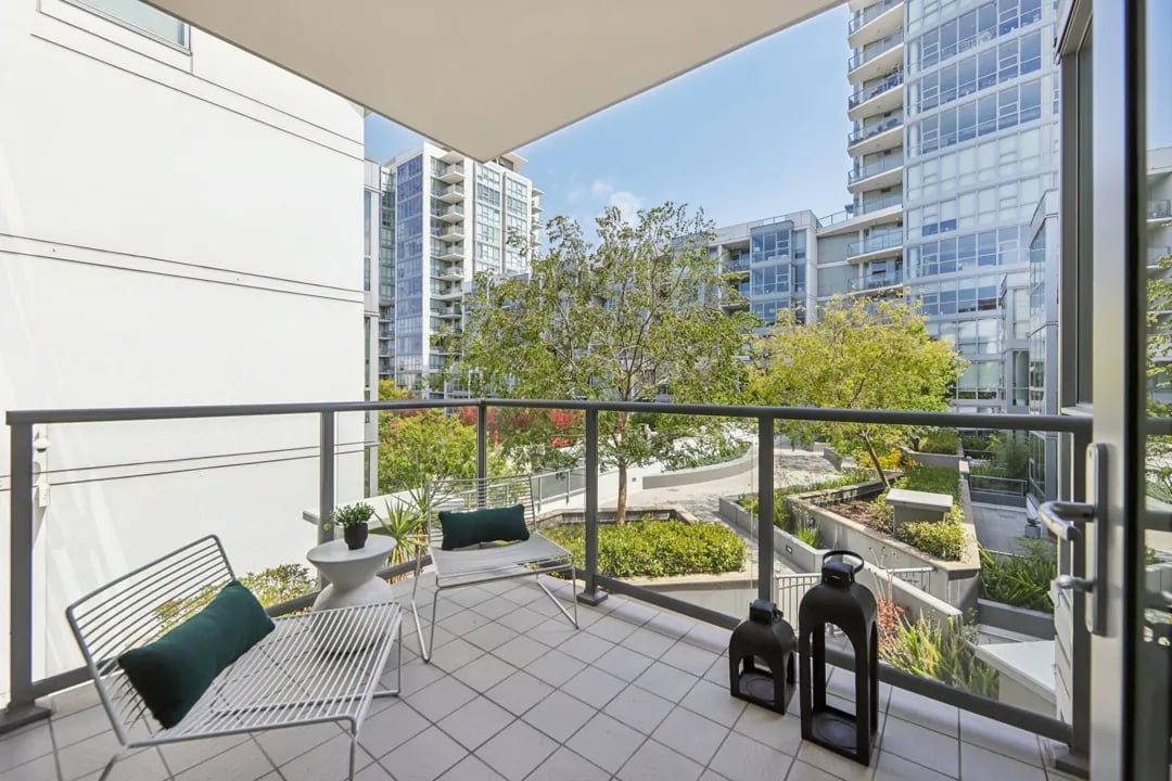 A bright and inviting balcony overlooks a lush courtyard garden surrounded by modern apartment buildings. 