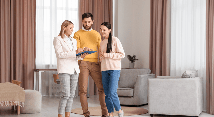 Three individuals stand in a living room, engaged with a tablet, surrounded by comfortable furniture and decor.