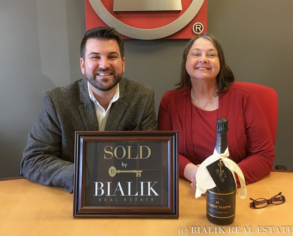 A smiling man and woman pose with a Bialik Real Estate sold sign and champagne.