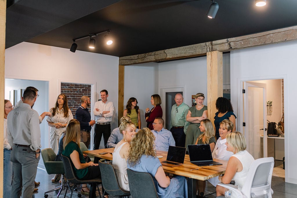 Adults gather around a wooden table with laptops in a modern office meeting room.