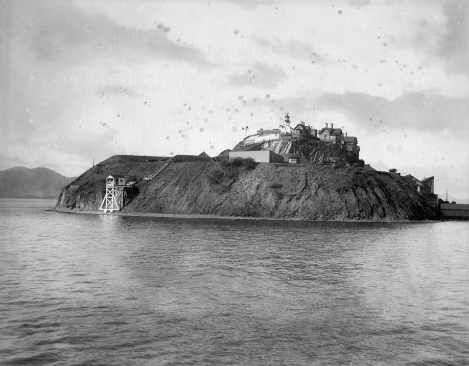 Black-and-white photograph of Alcatraz Island in San Francisco Bay, showing the rocky island with the prison buildings on top, a lighthouse, and a water tower, surrounded by water with distant hills in the background.