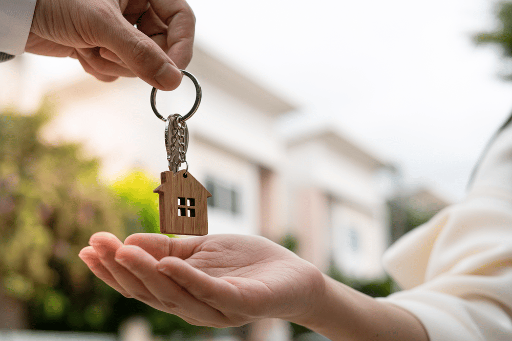 A hand holds a house-shaped keychain above an open palm, with a home blurred behind.