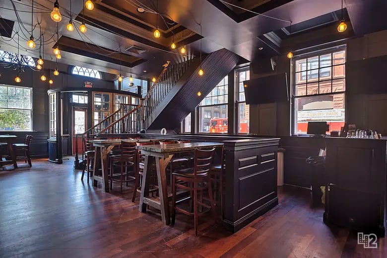 Restaurant interior with a long bar, wooden dining tables, an exposed staircase, and pendant lights.