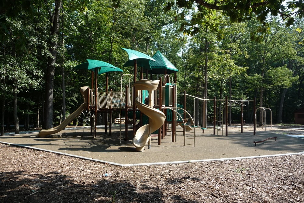 A playground with green roofs, slides, and climbing structures, surrounded by trees and mulch.