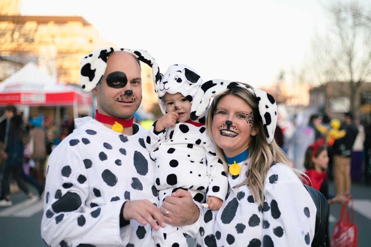 A family of three dressed as Dalmatian dogs poses for a Halloween photo, with other event-goers blurred in the background.