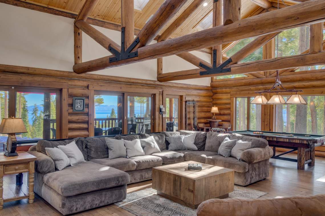 Log-cabin living room with vaulted timber beams, sectional sofa, and lake views through wide windows.