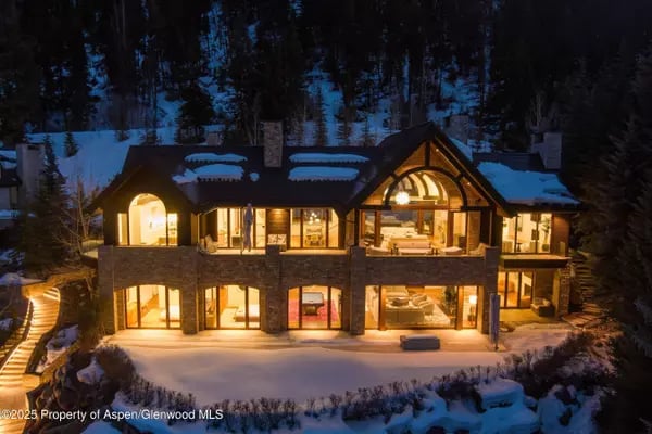 Two-story stone mountain home glowing warmly at night, surrounded by snow and evergreen forest.
