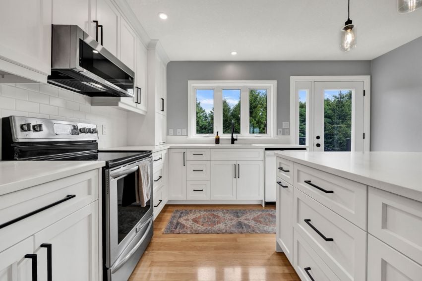 Bright white kitchen with a large island, stainless steel range, and windows overlooking trees.