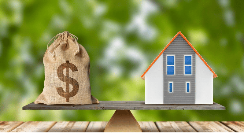 A money sack and model house balance on a wooden plank.