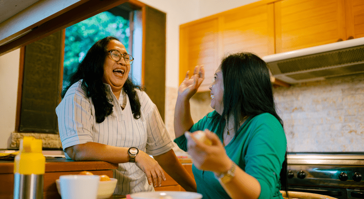 Two women are smiling and laughing in a kitchen. One leans against a counter, and the other gestures animatedly. The mood is joyful and warm.